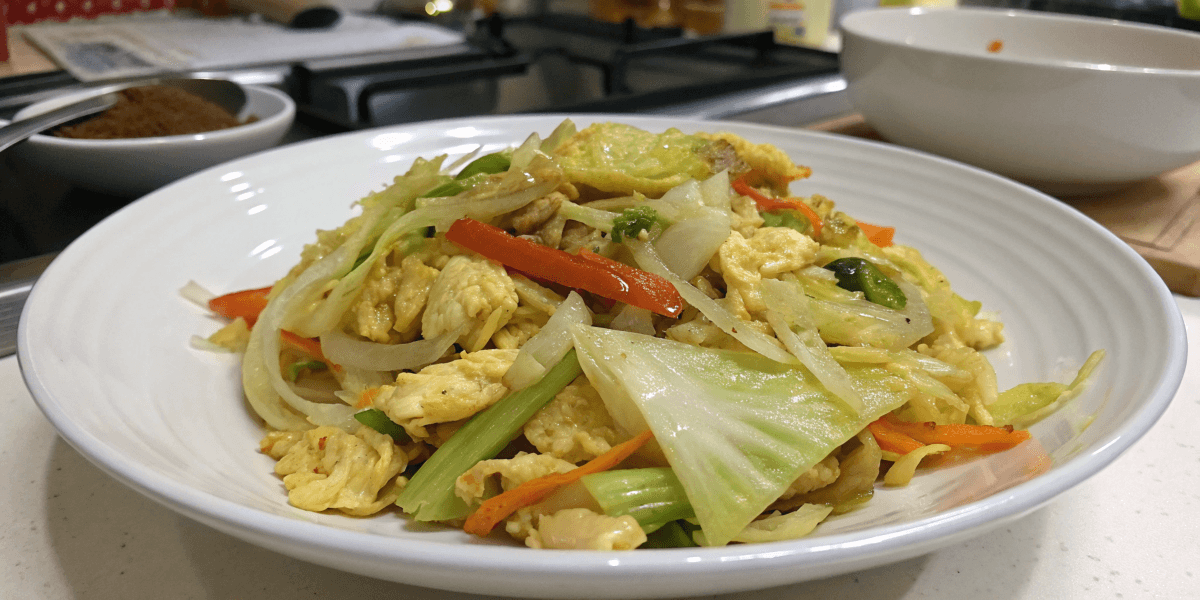 Close-up of a finished Cabbage and Egg recipe, garnished with sliced red bell peppers and chopped green onions. The stir-fry includes cabbage, scrambled eggs, onions, and other colorful vegetables, served on a white plate in a home kitchen setting